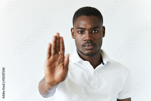 Headshot of dark-skinned young man wearing white polo shirt, preventing you from doing something with his open palm, looking at the camera with serious expression. Selective focus on the face