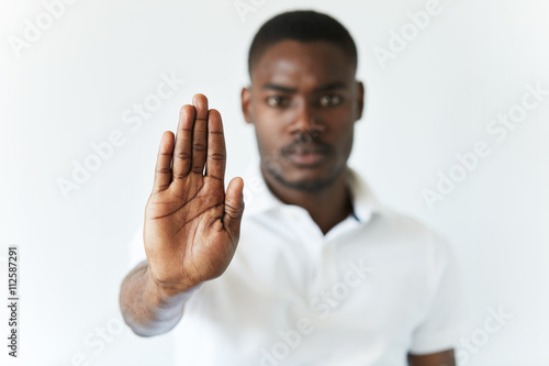 Portrait of serious African American male holding hand in stop sign, warning and preventing you from something bad, looking at the camera with worried expression. Selective focus on the palm
