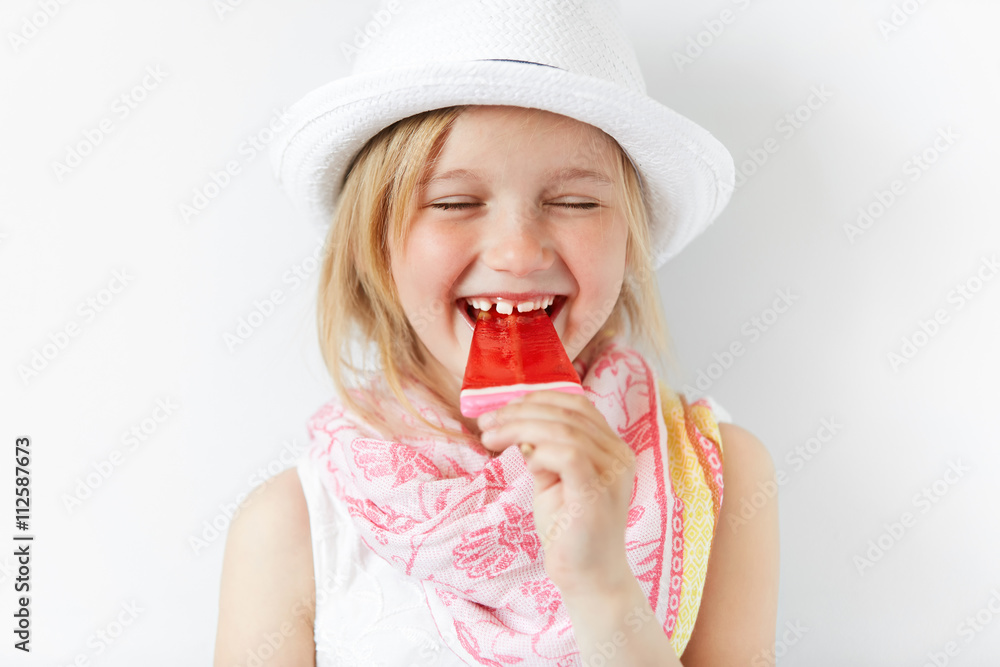 Laughing girl with red popsicle in white light room. Cheerful child eating her sugar candy with closed eyes. Delighted kid radiates positive emotions and summer holiday mood. Laughing girl with red popsicle in white light room. Cheerful child eating her sugar candy with closed eyes. Delighted kid radiates positive emotions and summer holiday mood.