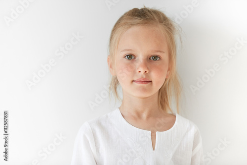 Little European girl in white children clothes looking peacefully at the camera indoors. Calm child standing quietly in restful manner with angelic look and innocent appearance. photo
