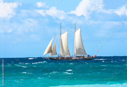 Vintage tall sail boat with masks high sailing off the coast on the blue choppy ocean lit up by sunshine.