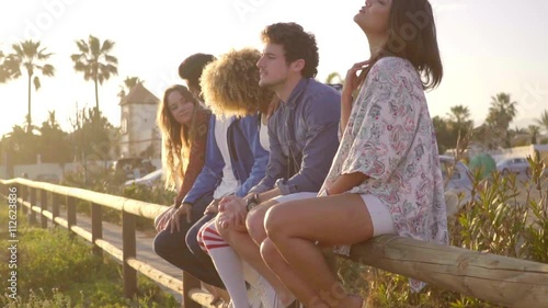 Group of young people sitting on wooden fence at the evening and make a gesture with the hands to encourage someone to come nearer and rejoicing. photo