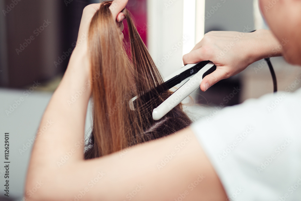 Fototapeta premium Hairstylist using a curling iron of a female client in a hairdressing salon, closeup view