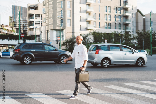 Young handsome business man holding briefcase walking on pedestrian crossing outdoor in the city, looking over smiling - business, work, happiness concept
