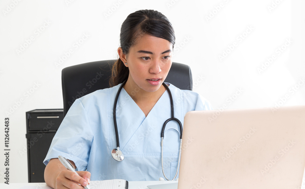 Young concentrated asian doctor with stethoscope around her neck looking at laptop and taking notes