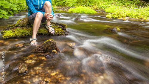 Fototapeta Naklejka Na Ścianę i Meble -  person sits on the stone covered with moss in the center of rapid flow of the river, holding his feet in clear water against the background of juicy greenery