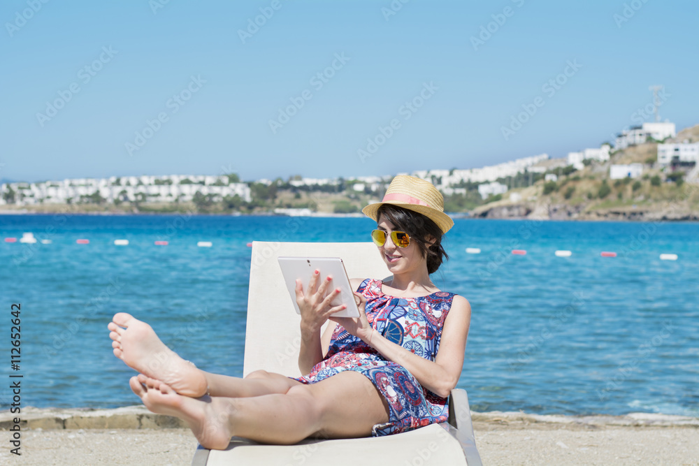Portrait of beautiful young woman sitting on a sunbed with tablet in the hands on a sea background.Summer holiday