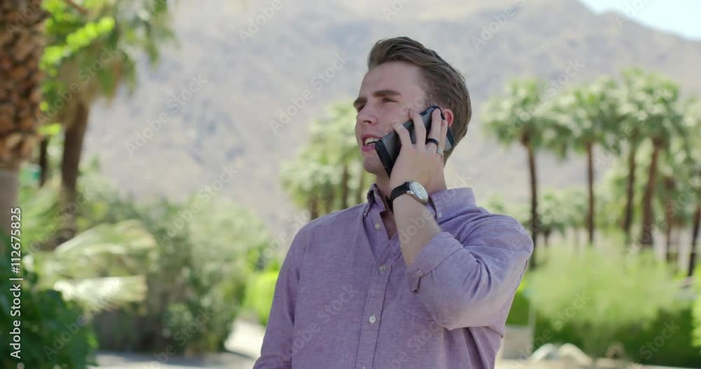 Handsome young man talking on a cellphone in Palm Springs, California. Background of palm trees and mountains. Close up, originally recorded hand held in 4K at 60fps Handsome young man talking on a cellphone in Palm Springs, California. Background of palm trees and mountains. Close up, originally recorded hand held in 4K at 60fps