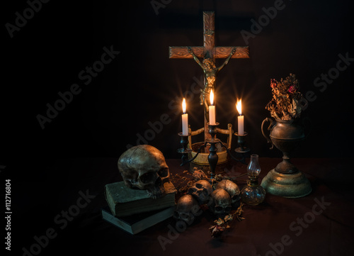 Low key of antique crafted crucifixion on crafted wooden cross with lit candles and skulls foreground