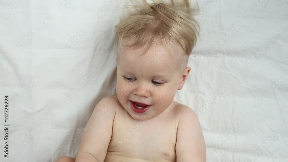 Adorable little baby is looking into the camera and is happy on a white bed sheet. The baby looks around and then smiles and laughs. Baby boy with blue eyes smiles at home Adorable little baby is looking into the camera and is happy on a white bed sheet. The baby looks around and then smiles and laughs. Baby boy with blue eyes smiles at home