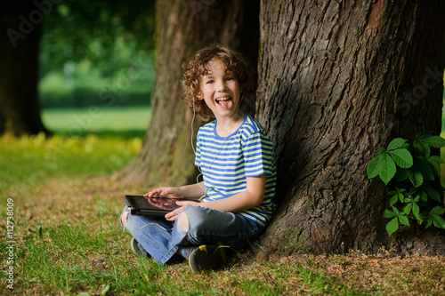 The cheerful boy sits under a tree with tablet on lap and puts out the tongue.