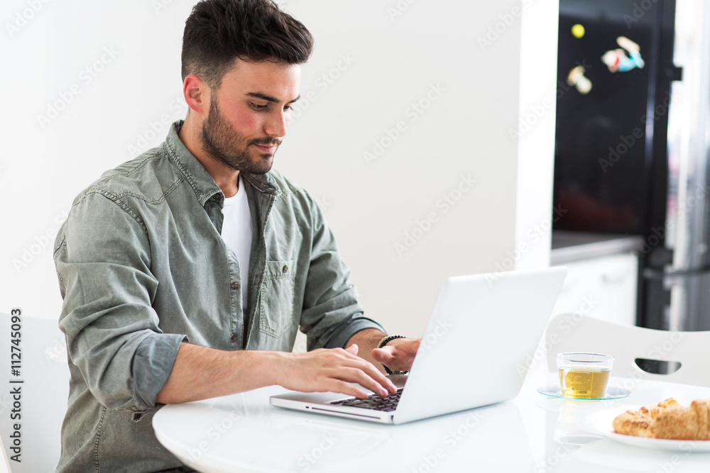 Young man doing something on laptop in his kitchen. Young man doing something on laptop in his kitchen.