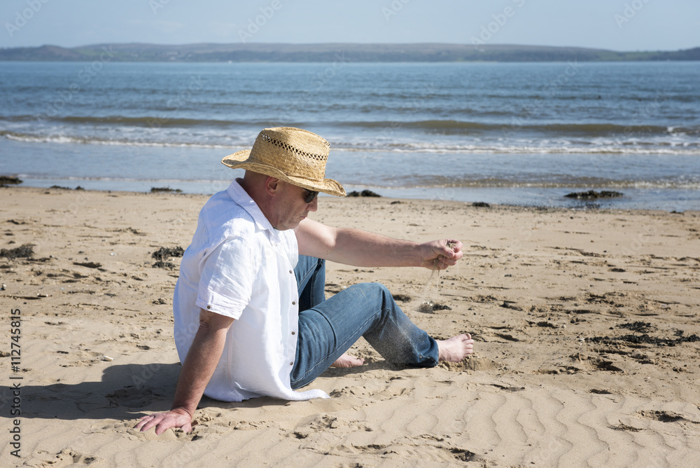 Mature man wearing casual clothes and a straw hat, playing with sand on a beach in summer.