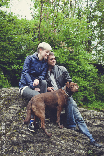 Happy family couple with dog hiking travelling in the forest. Beautiful sporty woman and young handsome man in leather jacket and cute pedigree dog together outdoors in the forest mountains near river