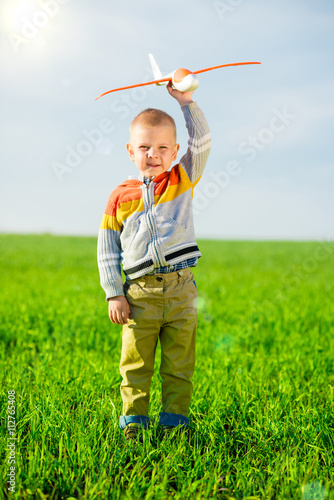 Happy kid playing with toy airplane against blue summer sky background. Boy throw foam plane in green field. Best childhood concept.