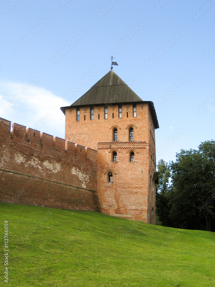 The tower and part of the walls of the ancient Kremlin in Veliky Novgorod, made of red bricks, located on a green hill.