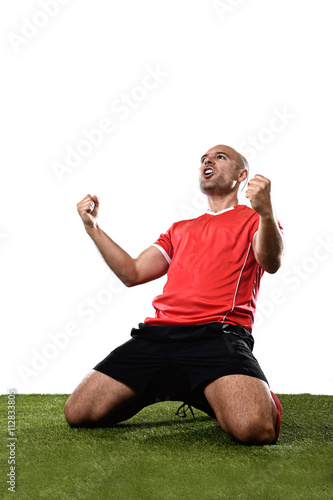 happy and excited football player in red jersey celebrating scoring goal kneeling on grass pitch