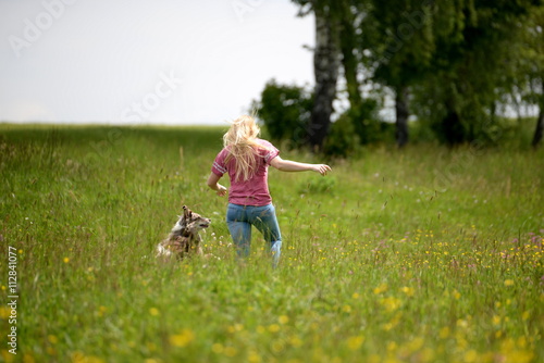 Wettlauf durch die Blumenwiese, Hütehund läuft mit blonder Frau durch Blumen