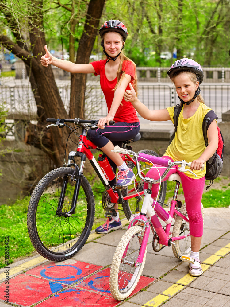 Fototapeta premium Bikes bicyclist girl. Girls wearing bicycle helmet with rucksack ciclyng bicycle show thumb up. Girls children cycling on yellow bike lane. Bike share program save money and time.