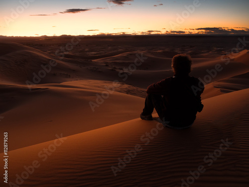 man sitting on a dune in the desert while watching the sunset. Morocco.