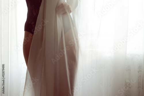 closeup on silhouette of hiding behind tulle curtain pretty lady with perfect fit body in black combies dress standing on light window copy space background