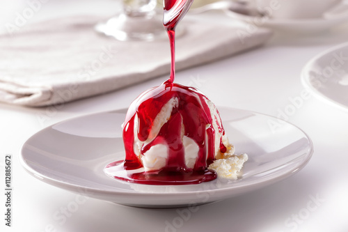 ice cream scoop with  pouring berry sauce from spoon on white plate close-up against decorated table

