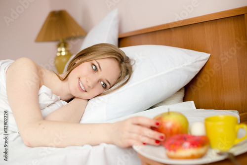 portrait of sexy blonde girl lying on white bed in lingerie and looking at plate with apple, donut & cup of hot drink on light copy space background photo