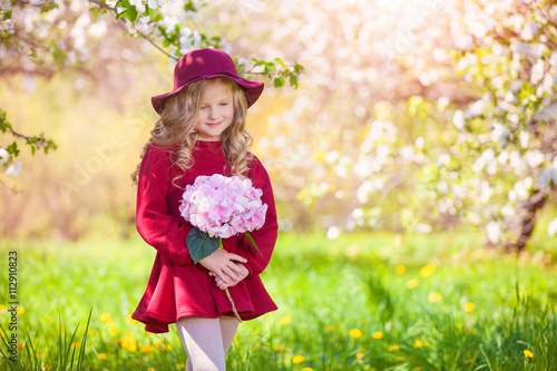 Beautiful little blonde girl in hat and burgundy dress. In her hand bouquet. Summer sunny day, lots of greenery and flowers