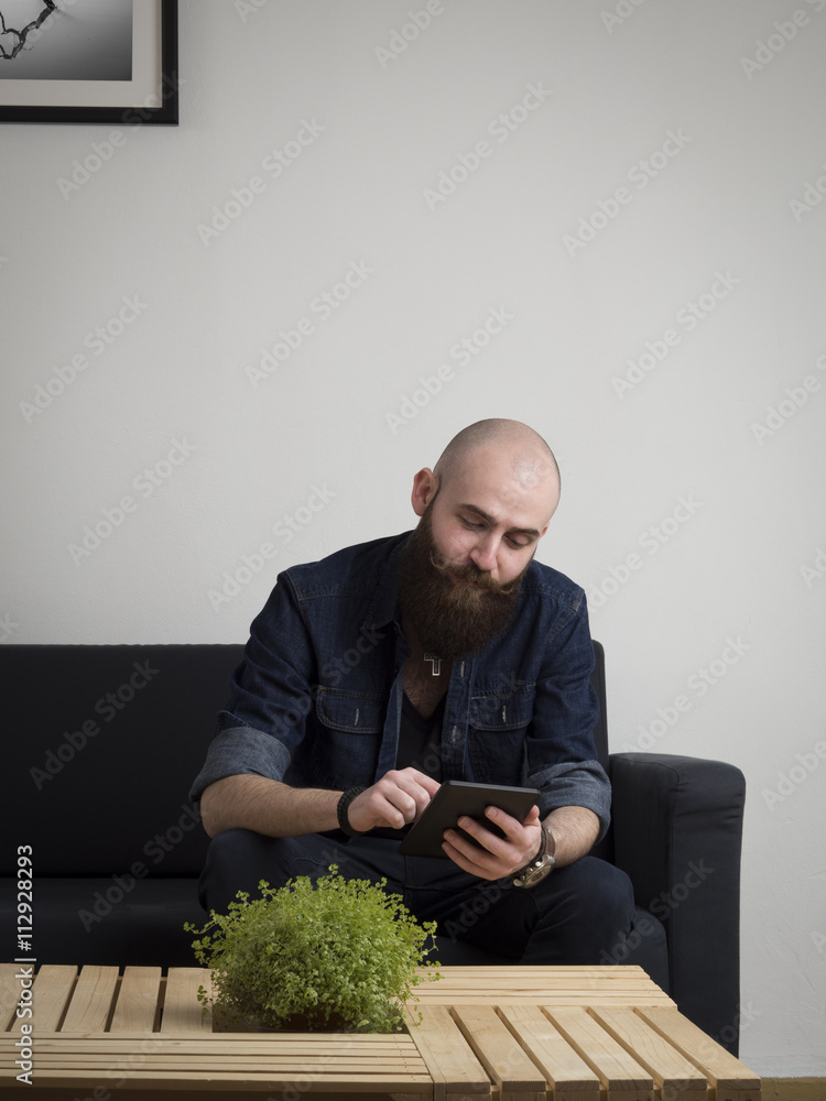 Young bearded hipster using digital tablet on the sofa at home. Young bearded hipster using digital tablet on the sofa at home.