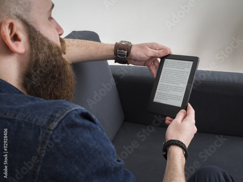 Young bearded man reading e book on the sofa. Shot from behind. photo
