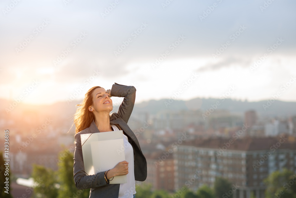 Young female entrepreneur enjoying business success against city and sunset background. Successful businesswoman smiling outdoor.