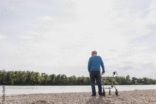 Back view of senior man standing with wheeled walker at riverside photo