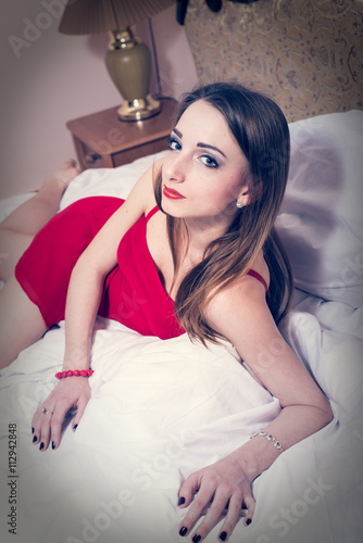 portrait of elegant sensual beautiful young lady having fun in red dress lying on the white bed on copy space bedroom background