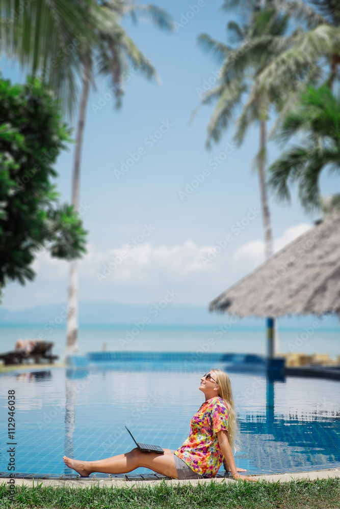Smiling freelancer sitting by pool near palm trees with laptop.