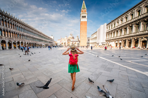 Young female traveler with hat and backpack standing on San Marco square with tower and basilica on the background in Venice. Back view with copy space