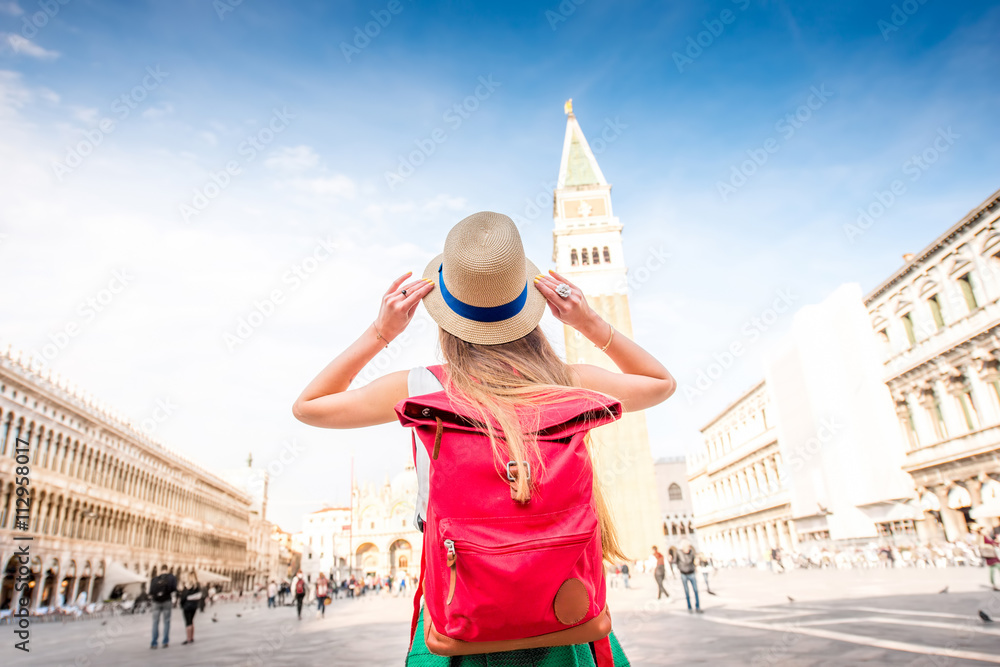 Young female traveler with hat and backpack standing on San Marco square with tower and basilica on the background in Venice. Back view - obrazy, fototapety, plakaty 