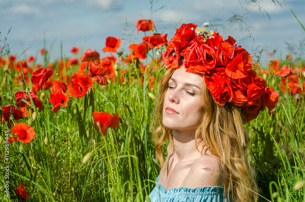 Naklejka premium Young beautiful happy girl with long hair in a white dress in the poppy field with a wreath on his head