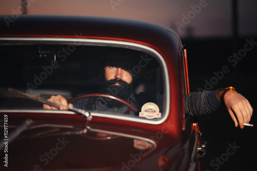 brutal bearded man with a smart mustache brunette sitting in a dark saloon car retro evening at sunset