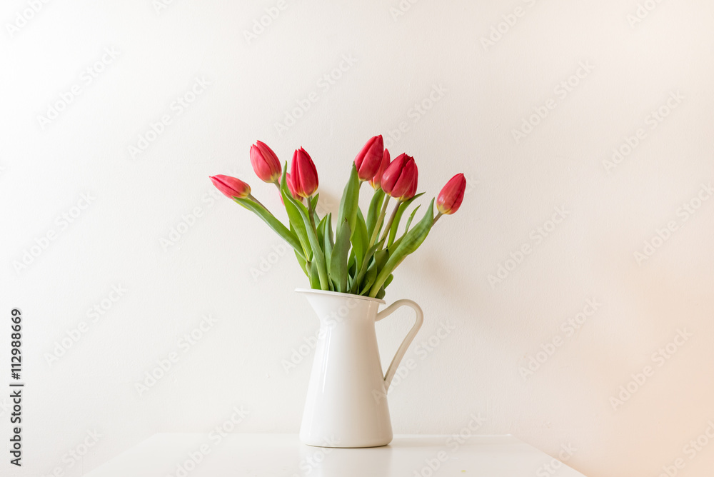 Red tulips in white jug on white table against white wall
