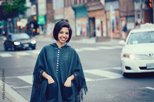Portrait of smiling young hipster latin hispanic girl woman with short hair bob, in torn jeans, grey blue poncho cape, standing outside in city street, looking in camera, street fashion concept © anoushkatoronto