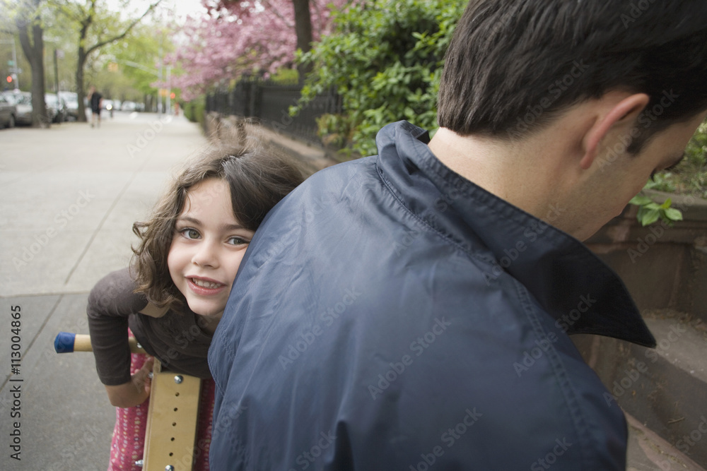 A young girl on a wooden scooter peeking around her father A young girl on a wooden scooter peeking around her father