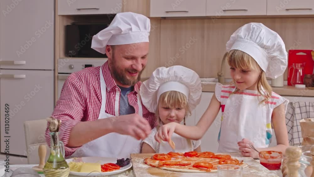 Dad giving slices of tomato and ham to his daughters and girls putting them on pizza dough Dad giving slices of tomato and ham to his daughters and girls putting them on pizza dough