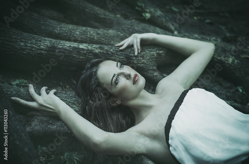 beautiful young woman in a long white dress with long wavy hair, lying on the large roots of a tree Fairy tale. Dream