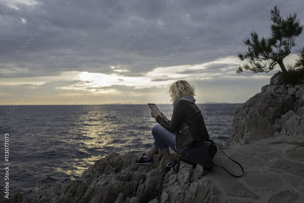 Beautiful blonde girl is seating on the stones near of the sea and watching the tablet