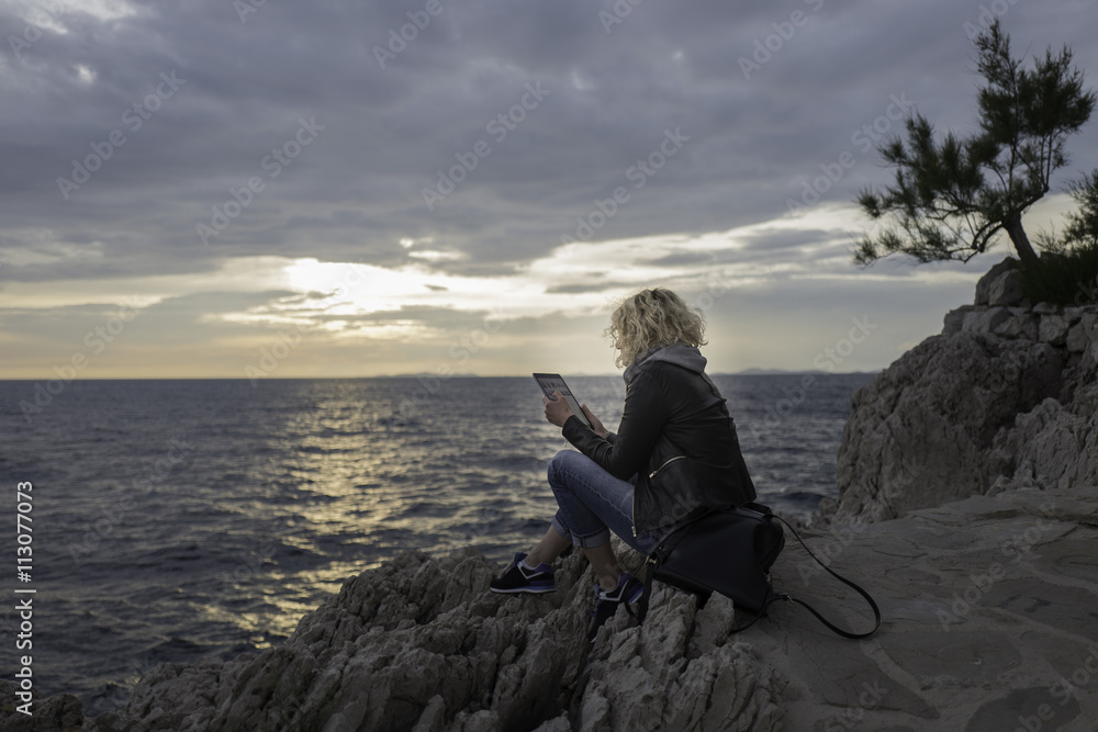 Beautiful blonde girl is seating on the stones near of the sea and watching the tablet Beautiful blonde girl is seating on the stones near of the sea and watching the tablet