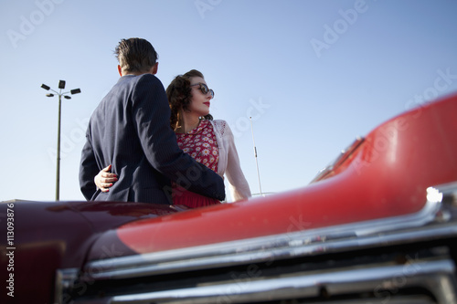 A cool, rockabilly couple with arms around each other by a vintage car