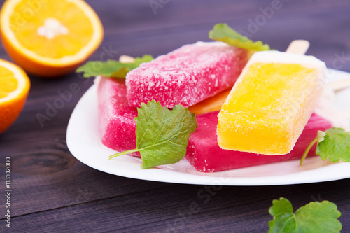 Colored frosty popsicles on a plate with mint and oranges on the wooden background