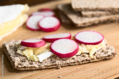 Wholemeal rye crispbread with brie cheese and radish slices, photographed with natural light (Selective Focus, Focus on the front of the first radish slice) photo