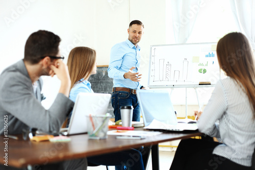 Handsome young man in glasses standing near whiteboard and pointing on the chart while his coworkers listening and sitting at the table