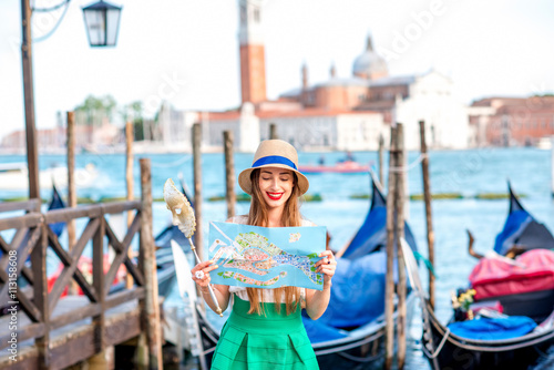 Young smiling woman traveling with paper map standing near San Marco square with gondolas on the background in Venice.